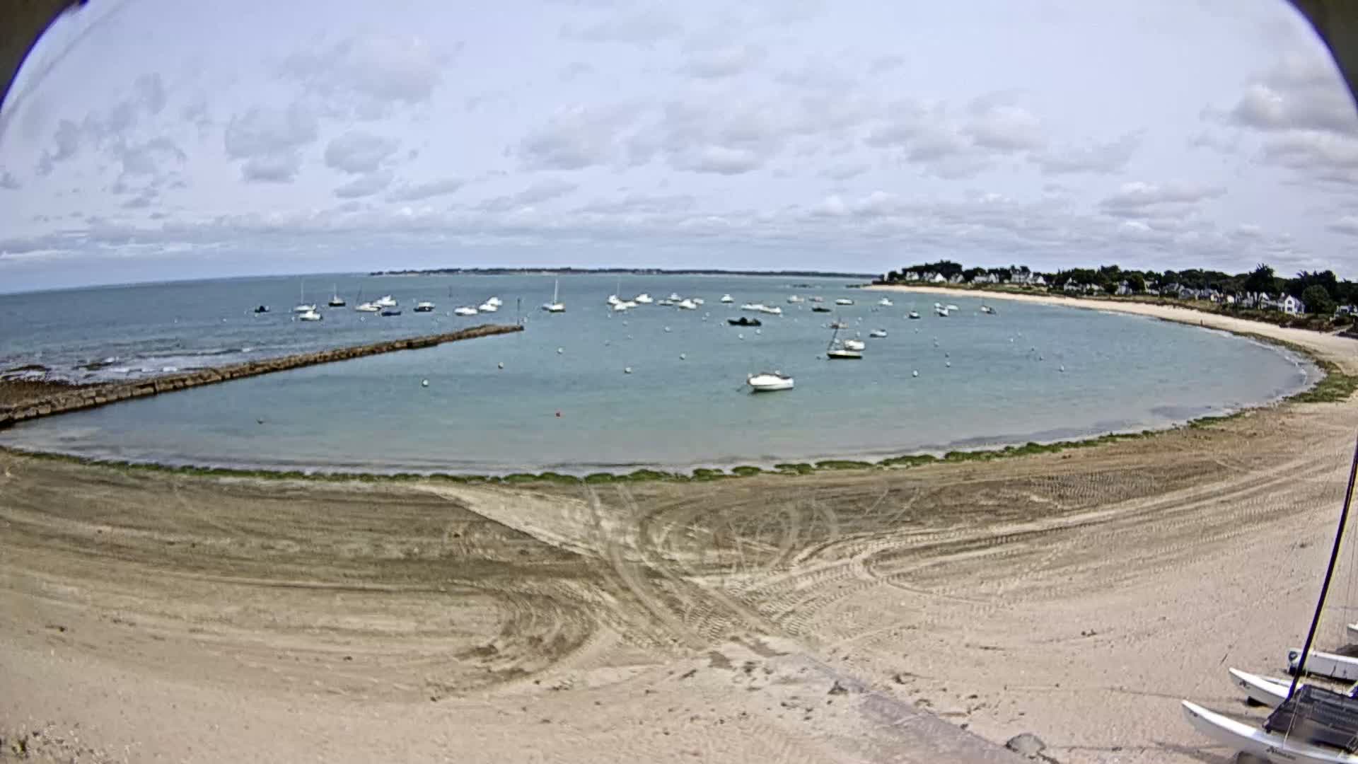 A sandy beach curves around a calm bay dotted with sailboats under a partly cloudy sky.