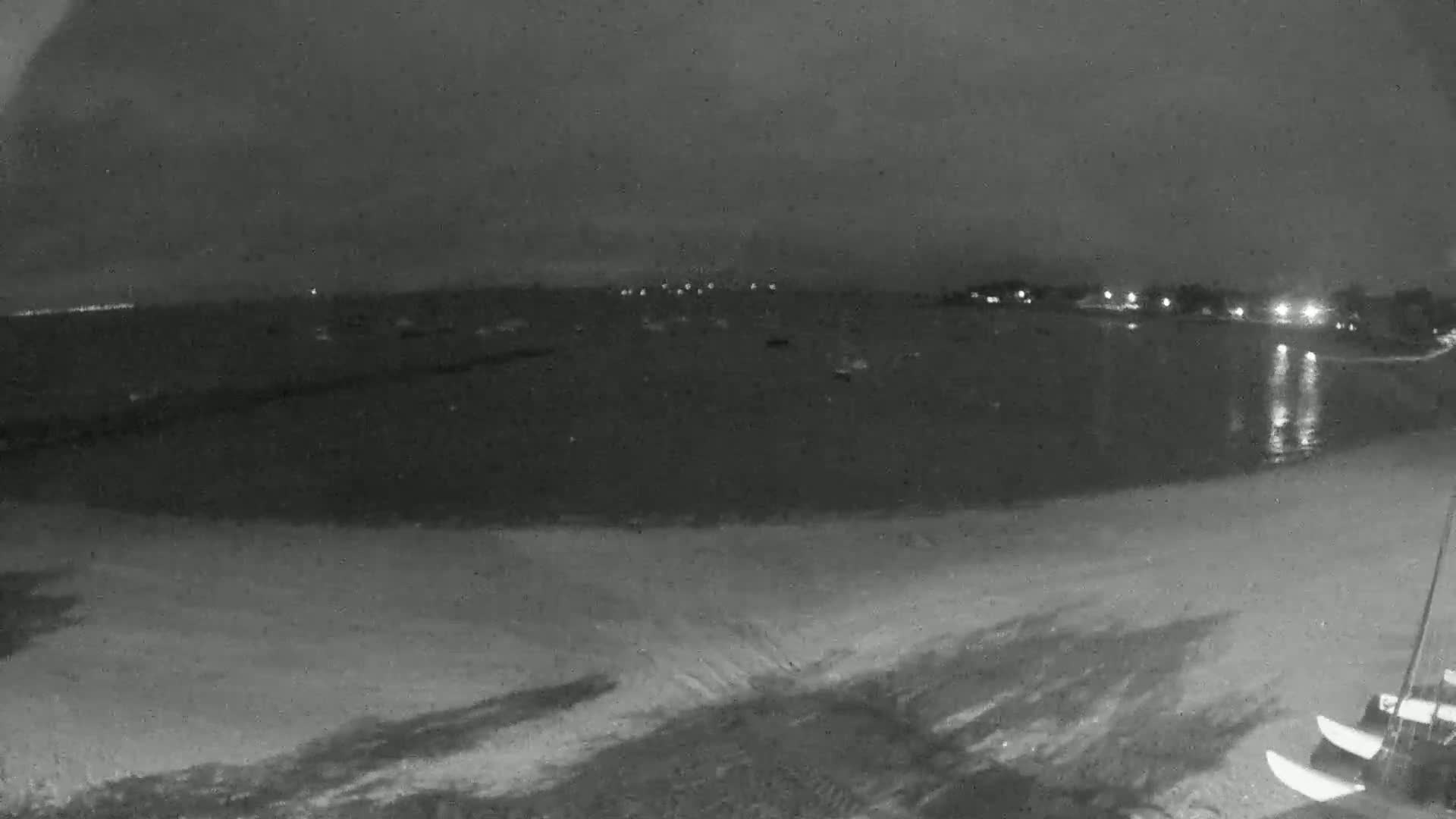 A nighttime view of a sandy beach, calm water with lights across the bay, and some boats on the shore.