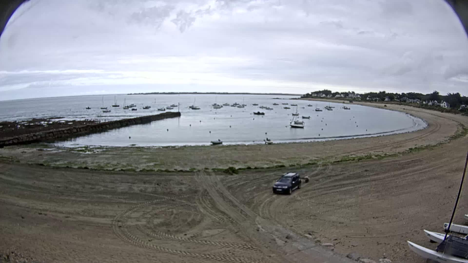 A sandy cove with several boats in the calm water is overlooked by a small town under a cloudy sky.