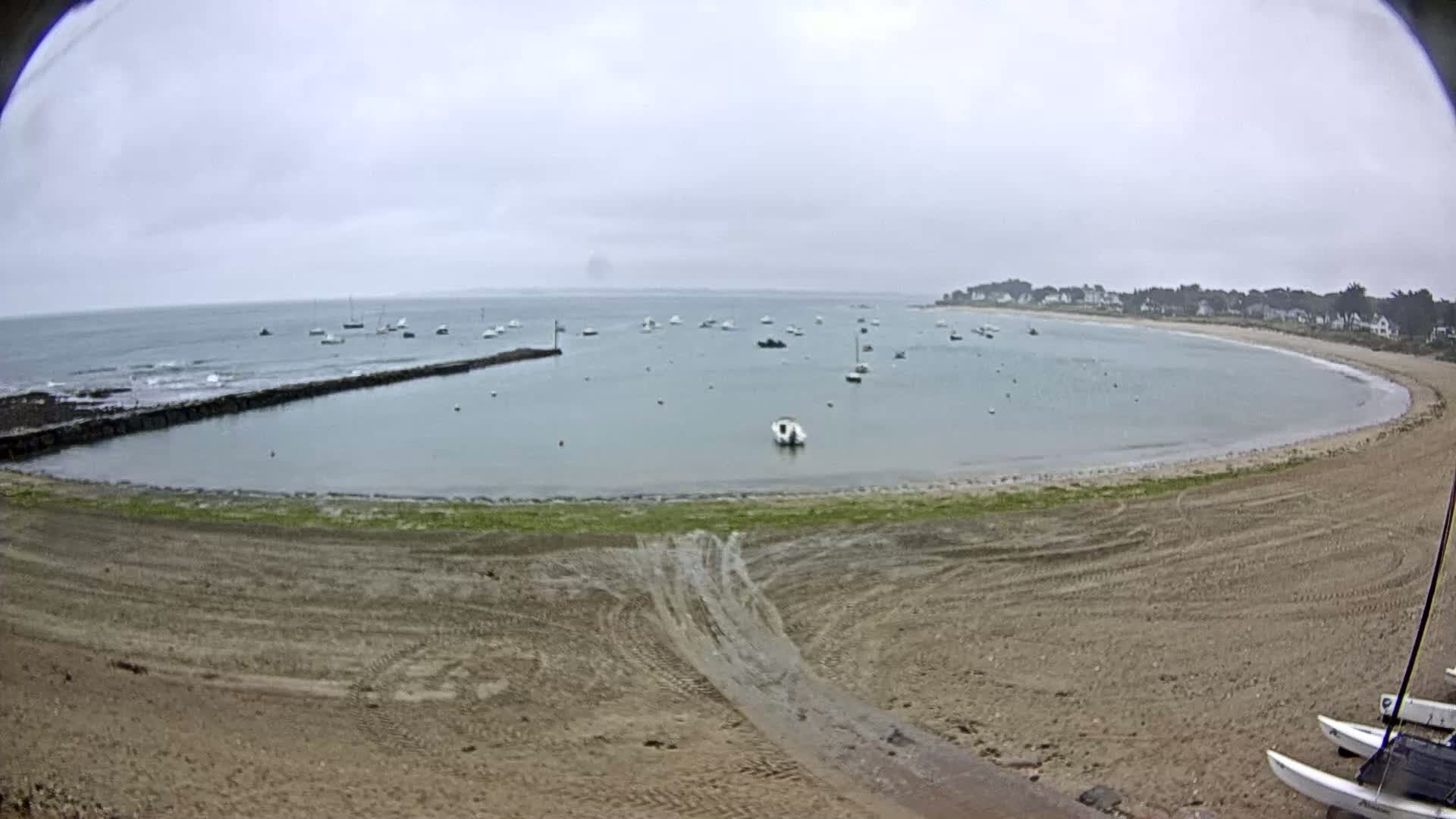 A sandy beach curves around a calm bay dotted with several small boats under an overcast sky.