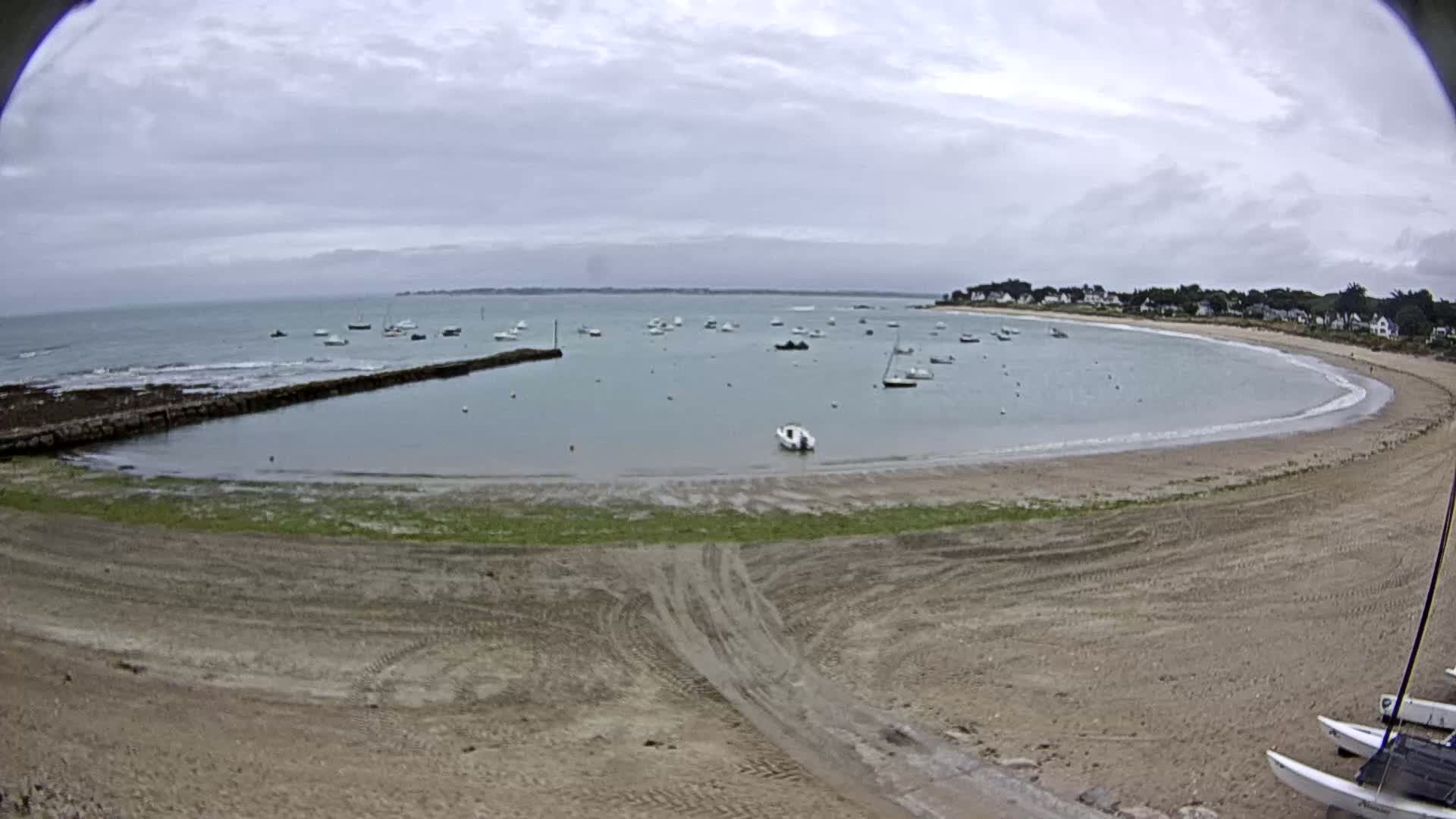 A sandy beach curves around a calm bay dotted with small boats under an overcast sky.