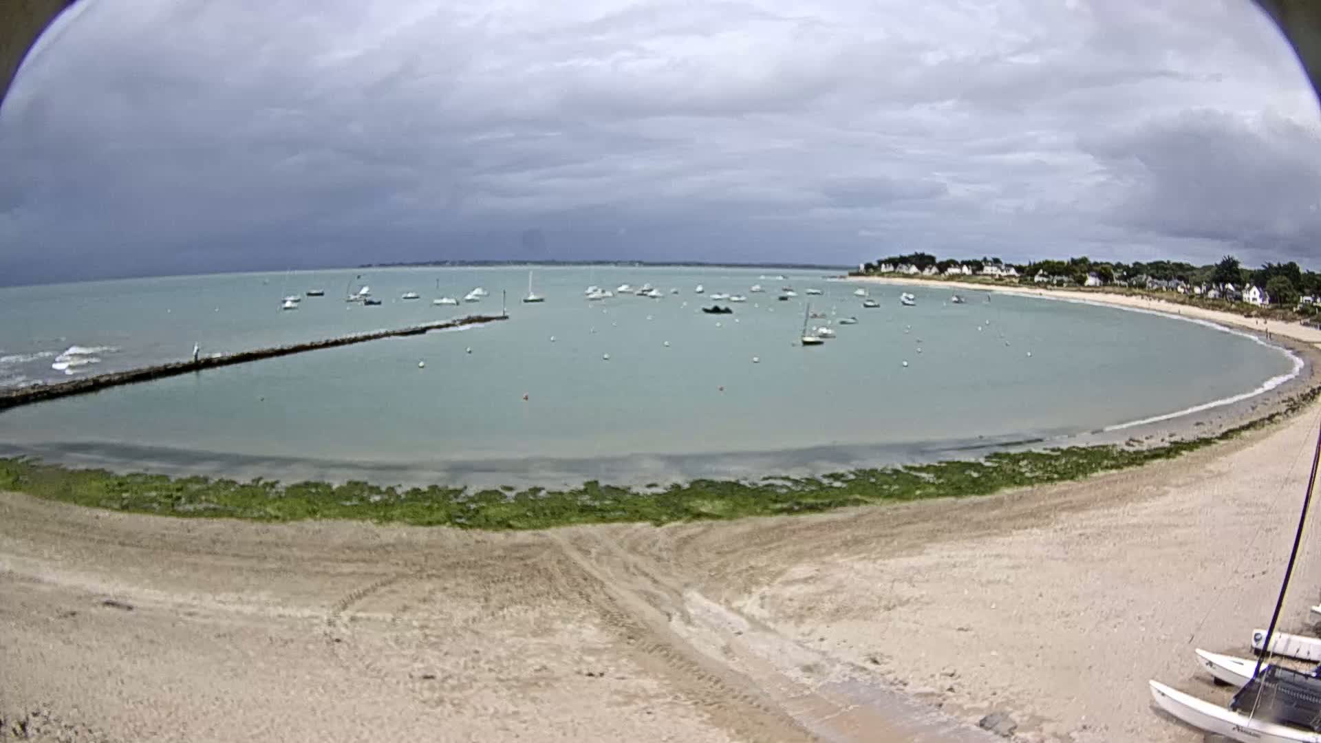 A sandy beach curves around a calm, light-green bay dotted with numerous boats under a cloudy sky.
