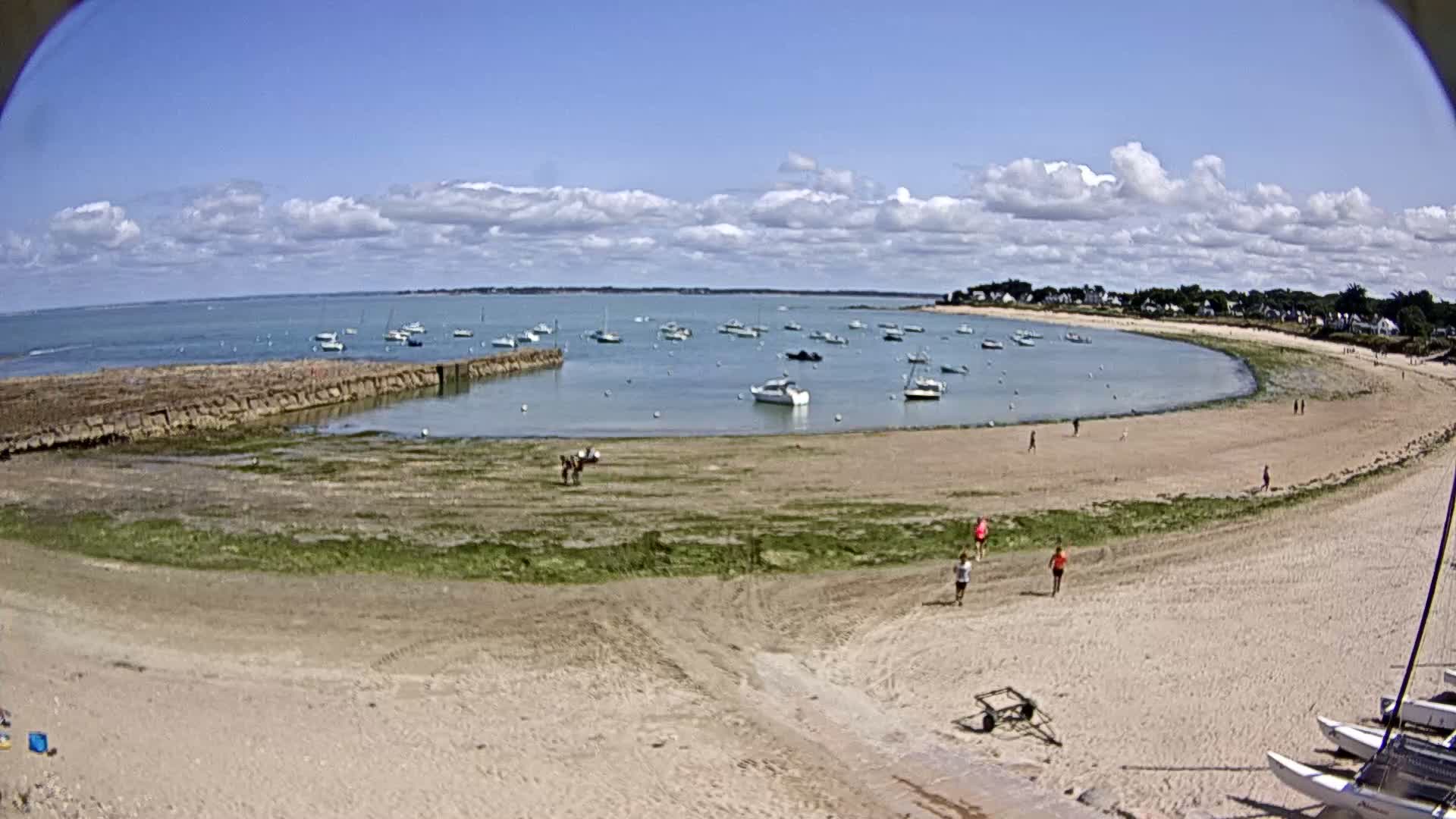A sandy beach curves around a calm bay filled with small boats under a partly cloudy blue sky.
