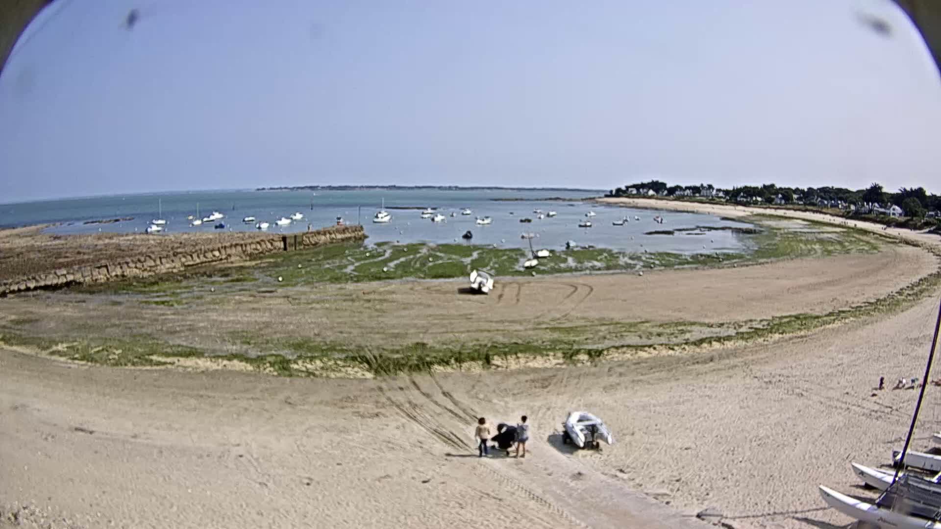 A sandy beach curves around a calm bay under a clear blue sky, with several boats in the water and a few people walking on the sand.