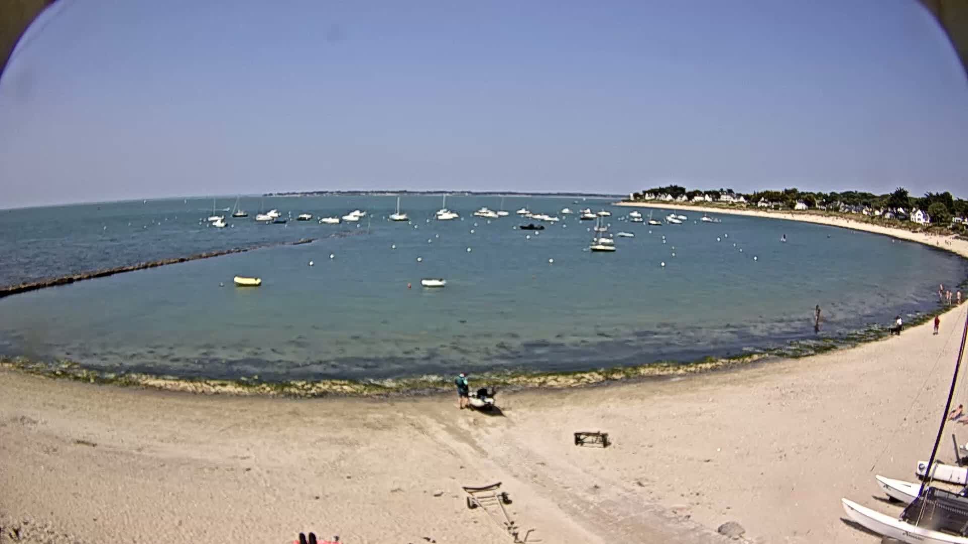 A sandy beach curves around a calm, clear bay with numerous boats at anchor on a sunny day.