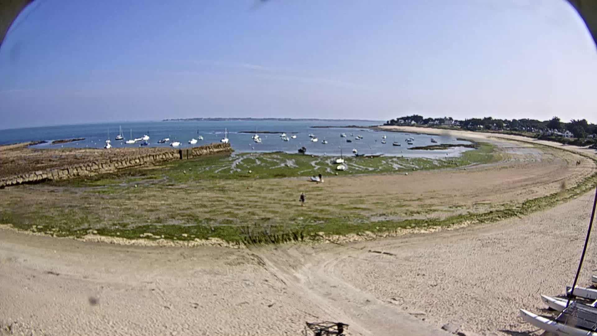 A sandy beach curves around a calm bay with several sailboats at anchor under a clear blue sky.