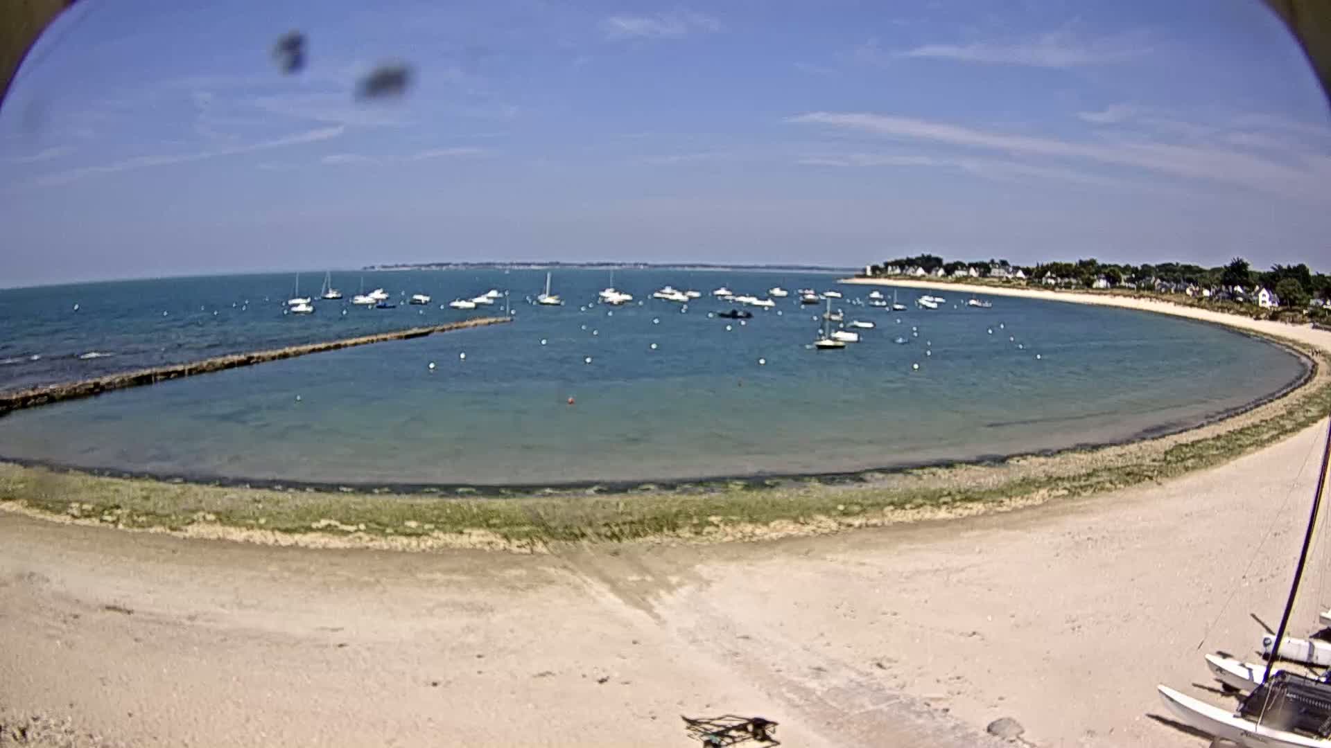 A sandy beach curves around a calm bay filled with sailboats under a sunny sky.