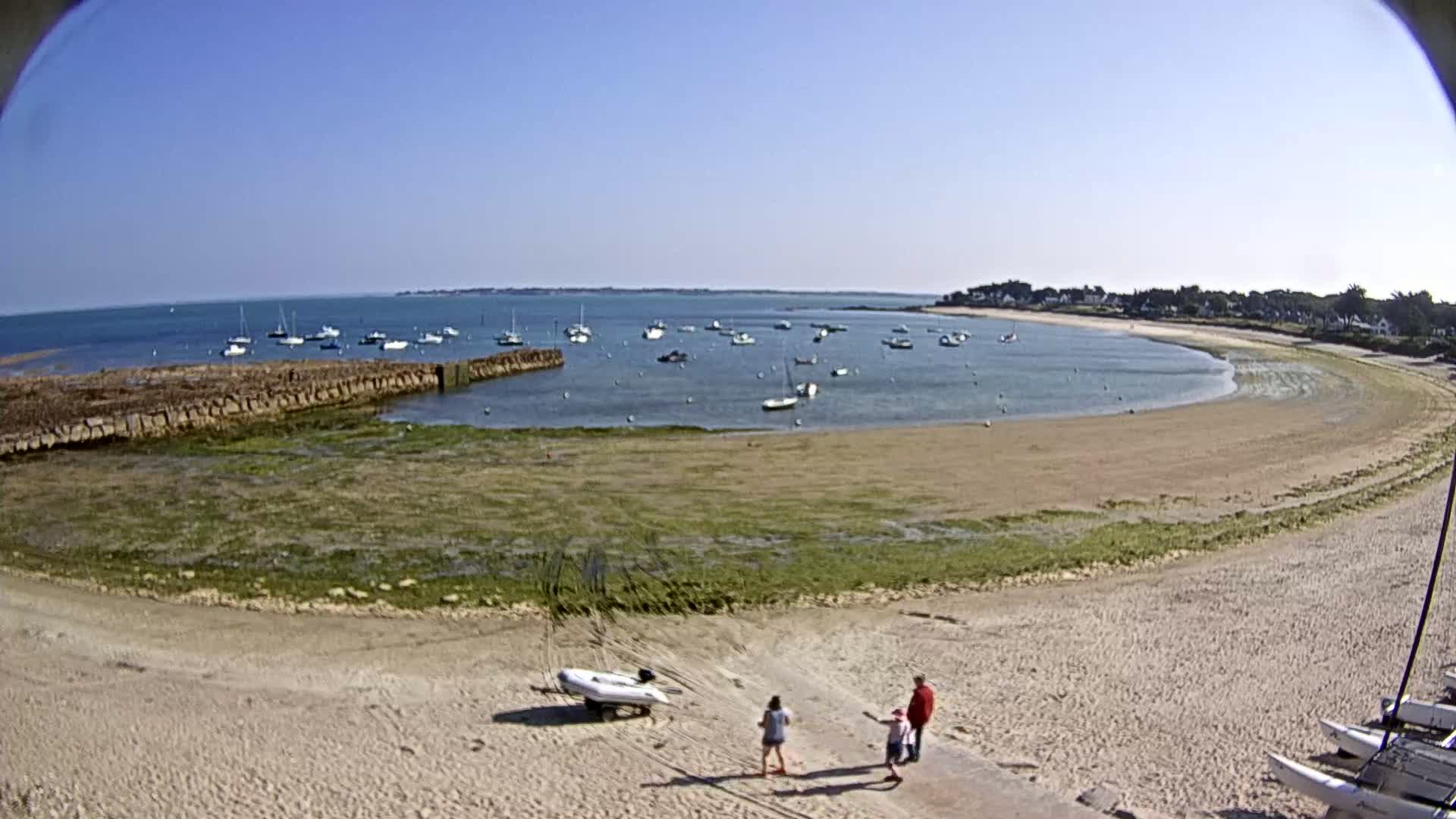 A sandy beach curves around a calm bay with numerous small boats anchored, under a clear blue sky.