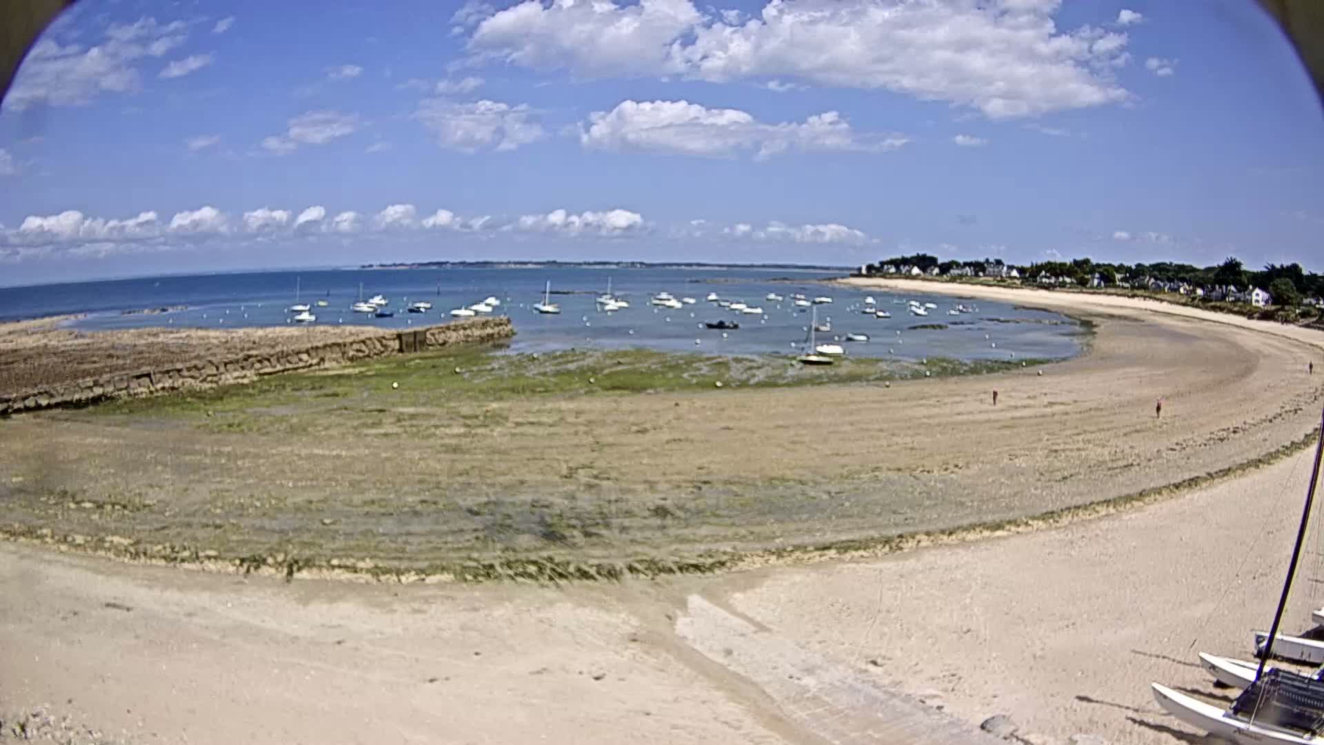 A sandy beach curves around a calm bay, dotted with numerous small boats under a partly cloudy blue sky.