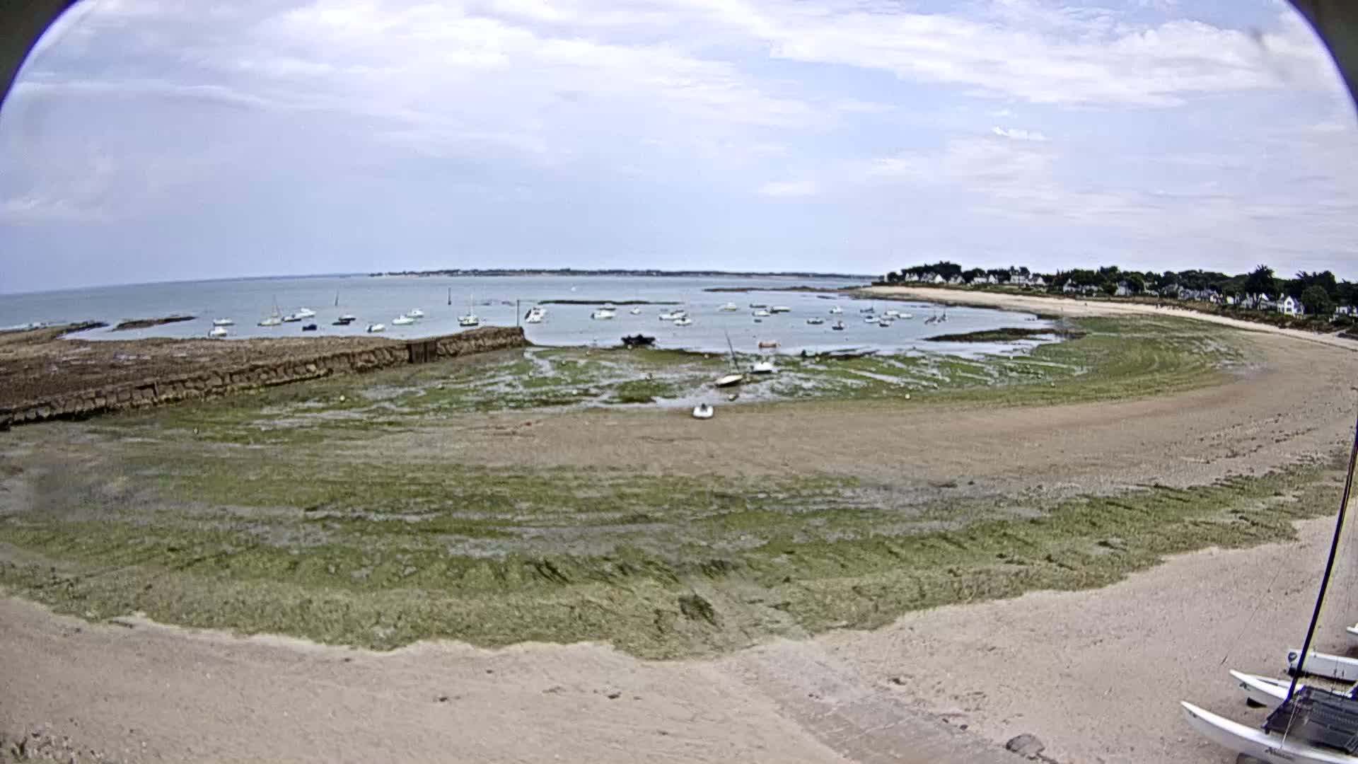 A mostly sunny day reveals a sandy beach with green algae, numerous small boats in shallow water, and a small town in the distance.