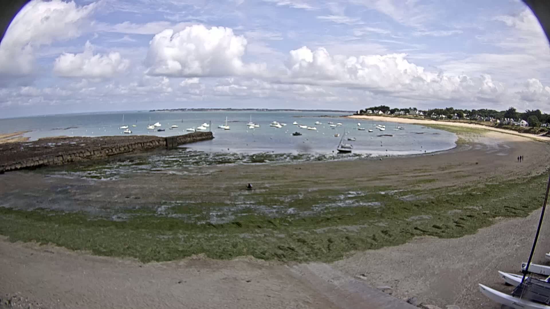 A partly sunny day shows a sandy cove with a few sailboats in the water, a stone wall, and seaweed-covered mudflats at low tide.