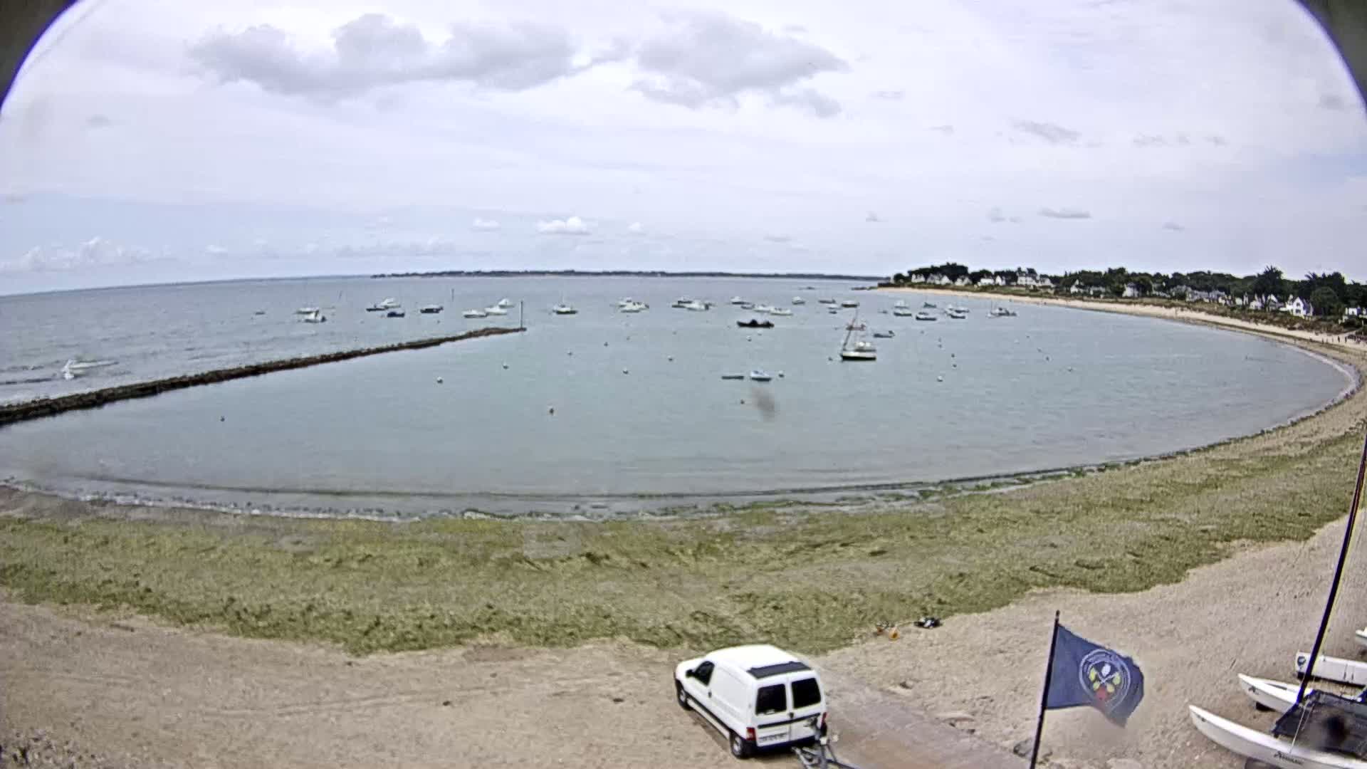 A sandy beach curves around a calm bay dotted with numerous small boats under a partly cloudy sky.