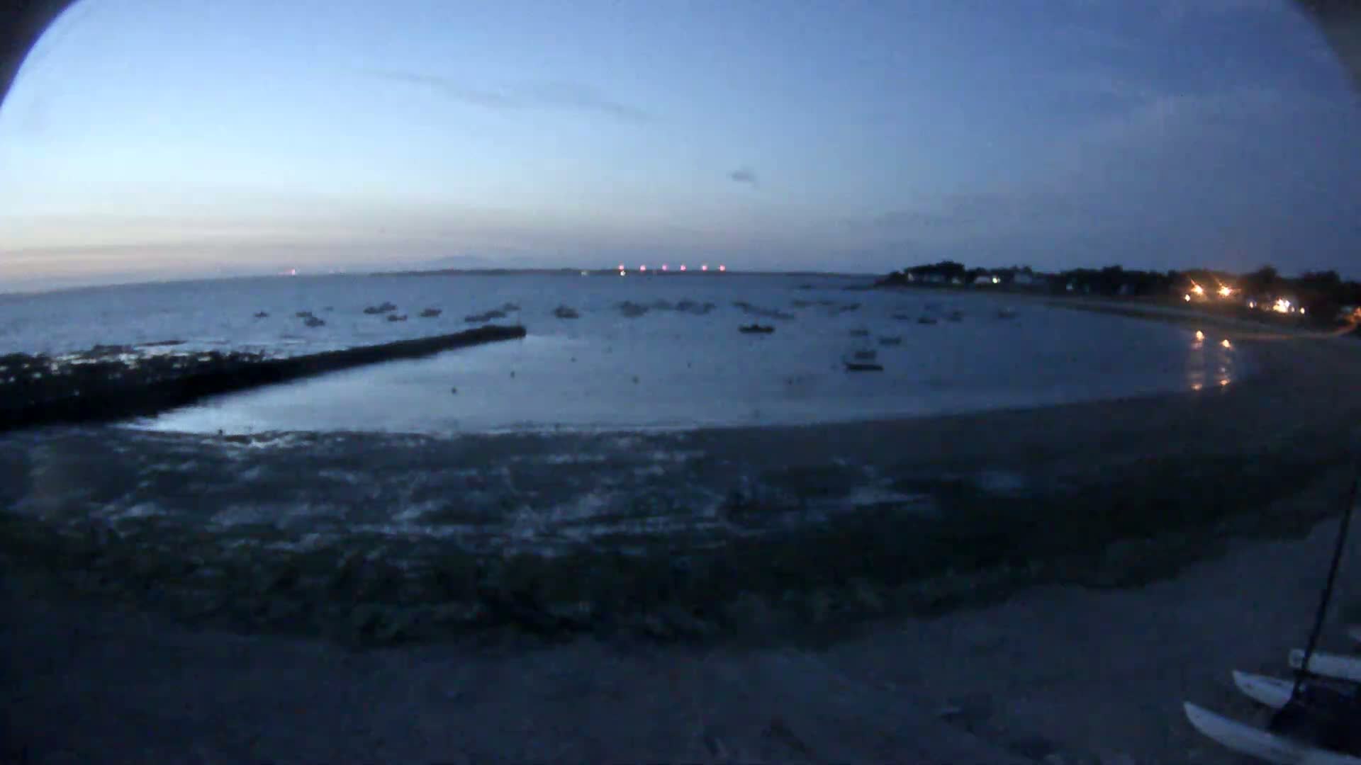 A sandy beach curves around a calm bay dotted with numerous small boats under a partly cloudy sky.