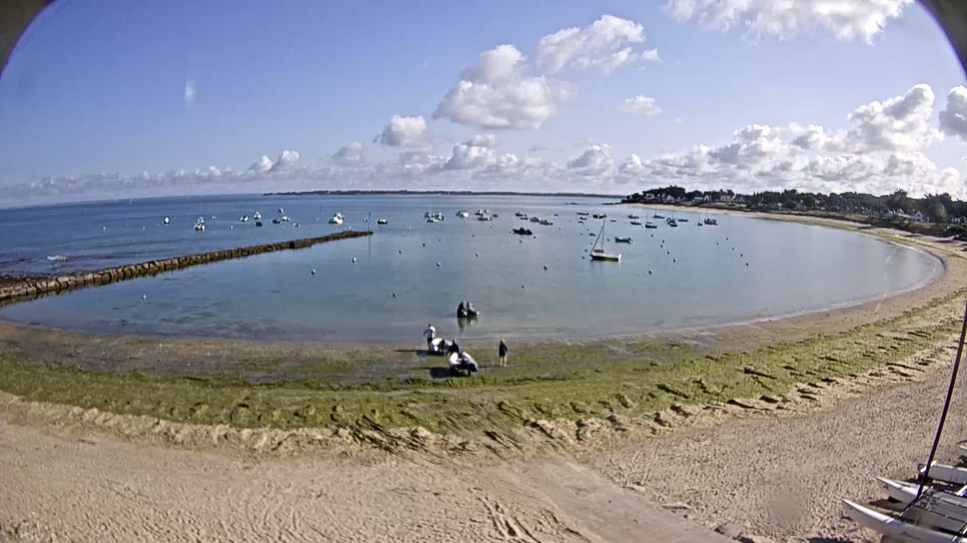A sandy beach curves around a calm, shallow bay filled with numerous small boats under a partly cloudy sky.