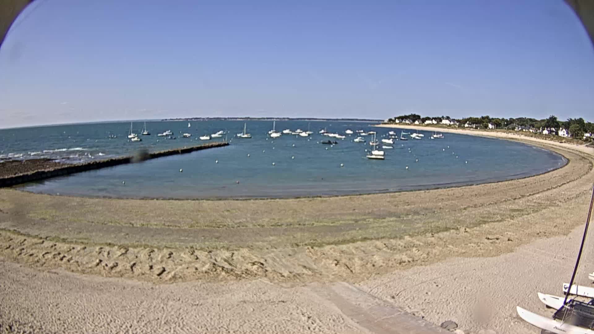 A sandy beach curves around a calm bay filled with sailboats under a clear blue sky.