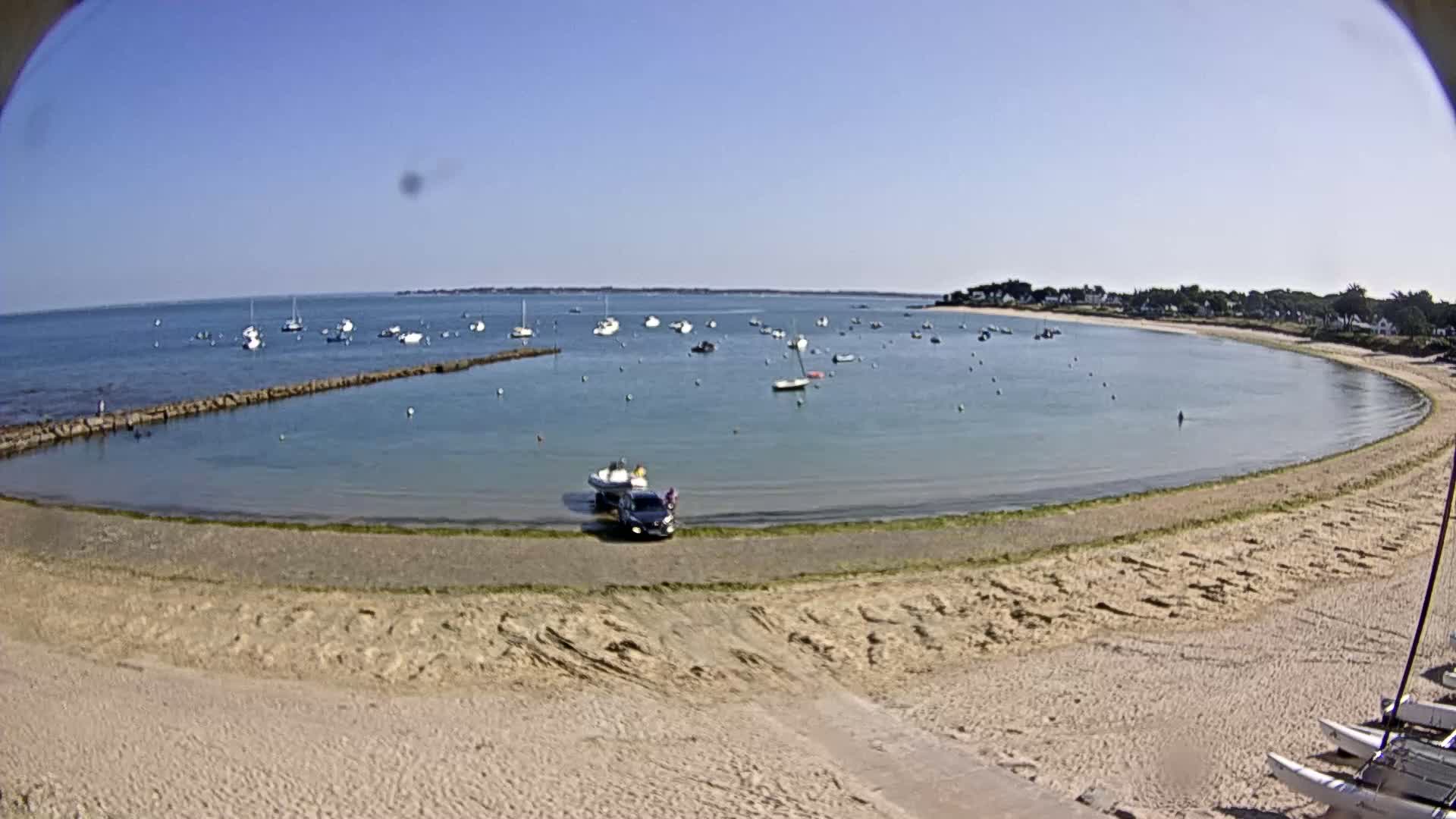 A sandy beach curves around a calm, shallow bay filled with small boats under a clear blue sky.