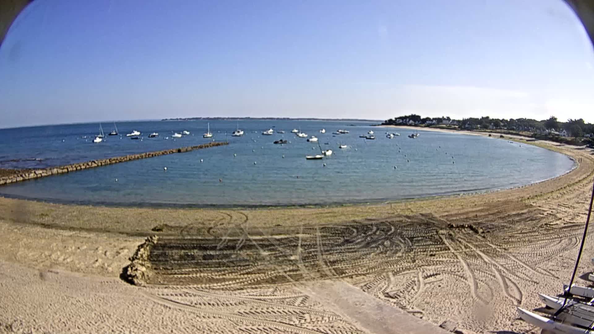 A sandy beach curves around a calm bay filled with numerous small boats under a clear blue sky.