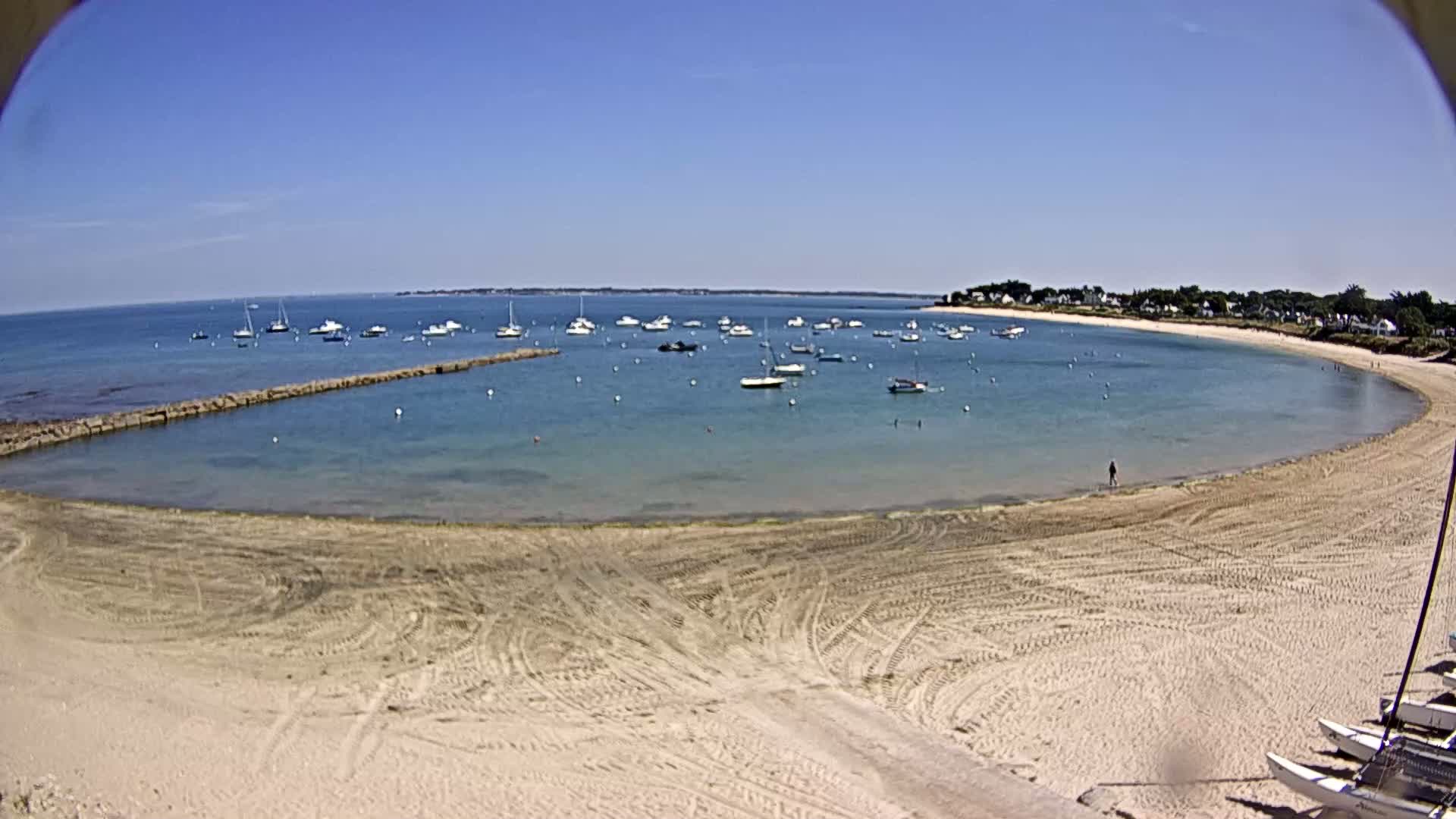 A sandy beach curves around a calm, clear bay filled with numerous small boats under a bright, sunny sky.