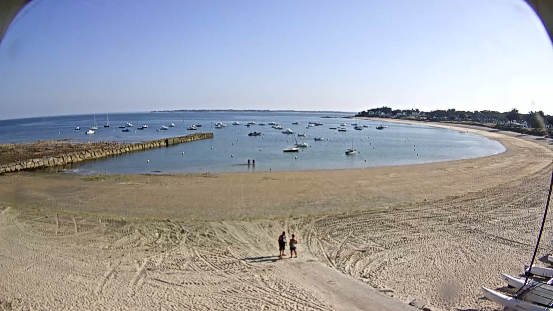 A sandy beach curves around a calm bay filled with numerous small boats under a clear blue sky.