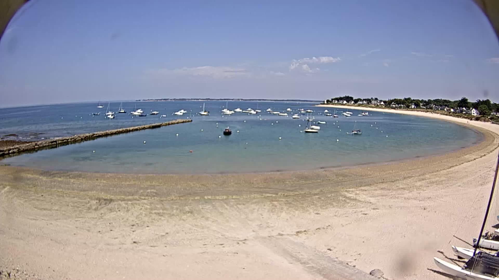 A sandy beach curves around a calm, clear bay dotted with sailboats under a sunny sky.