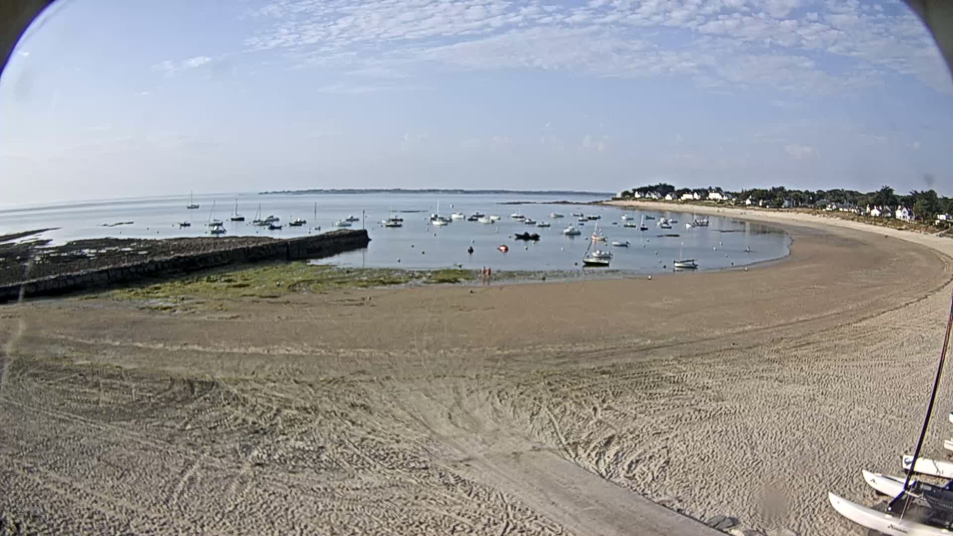 A sandy beach curves around a calm bay dotted with sailboats under a partly cloudy sky.