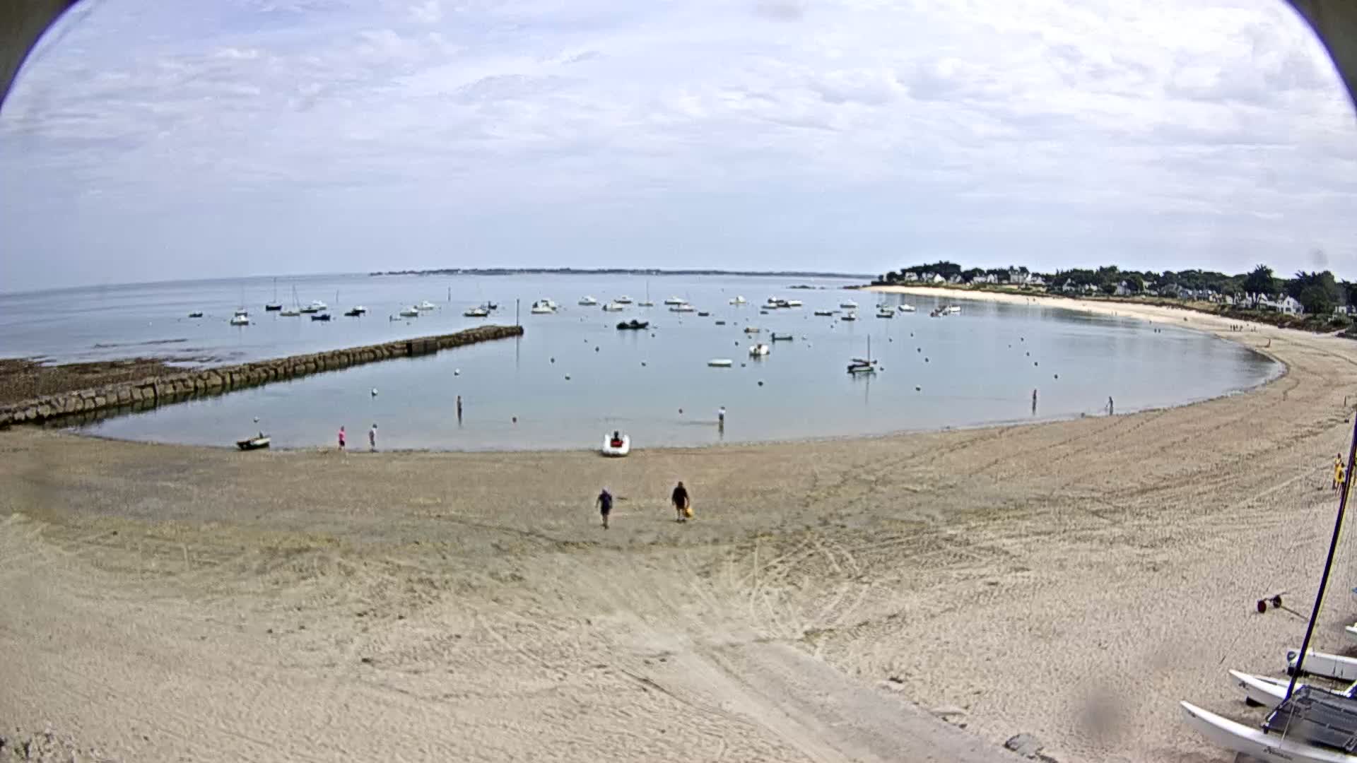A sandy beach curves around a calm bay filled with numerous small boats under a partly cloudy sky.