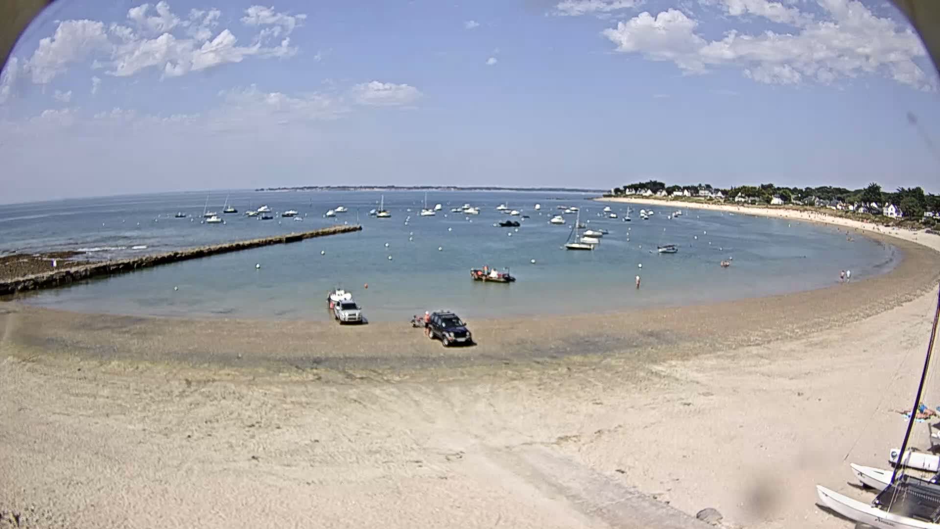 A sandy beach curves around a calm bay filled with numerous small boats under a partly cloudy sky.