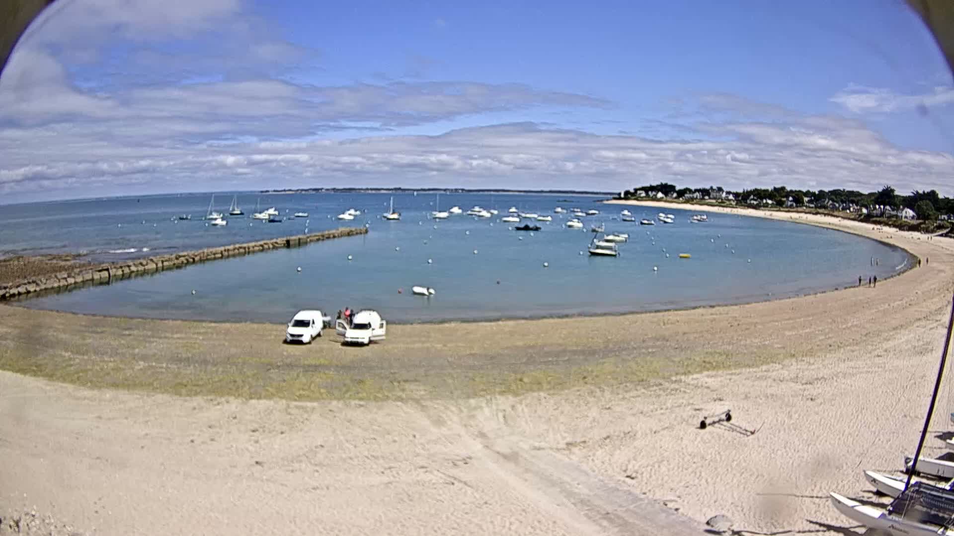 A sandy cove with several boats in the water and a few cars parked on the beach under a partly cloudy sky.