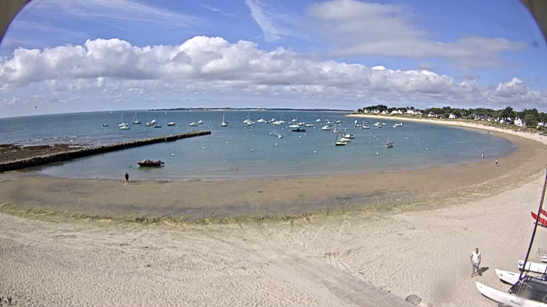 A sandy beach curves around a calm bay filled with sailboats under a partly cloudy sky.