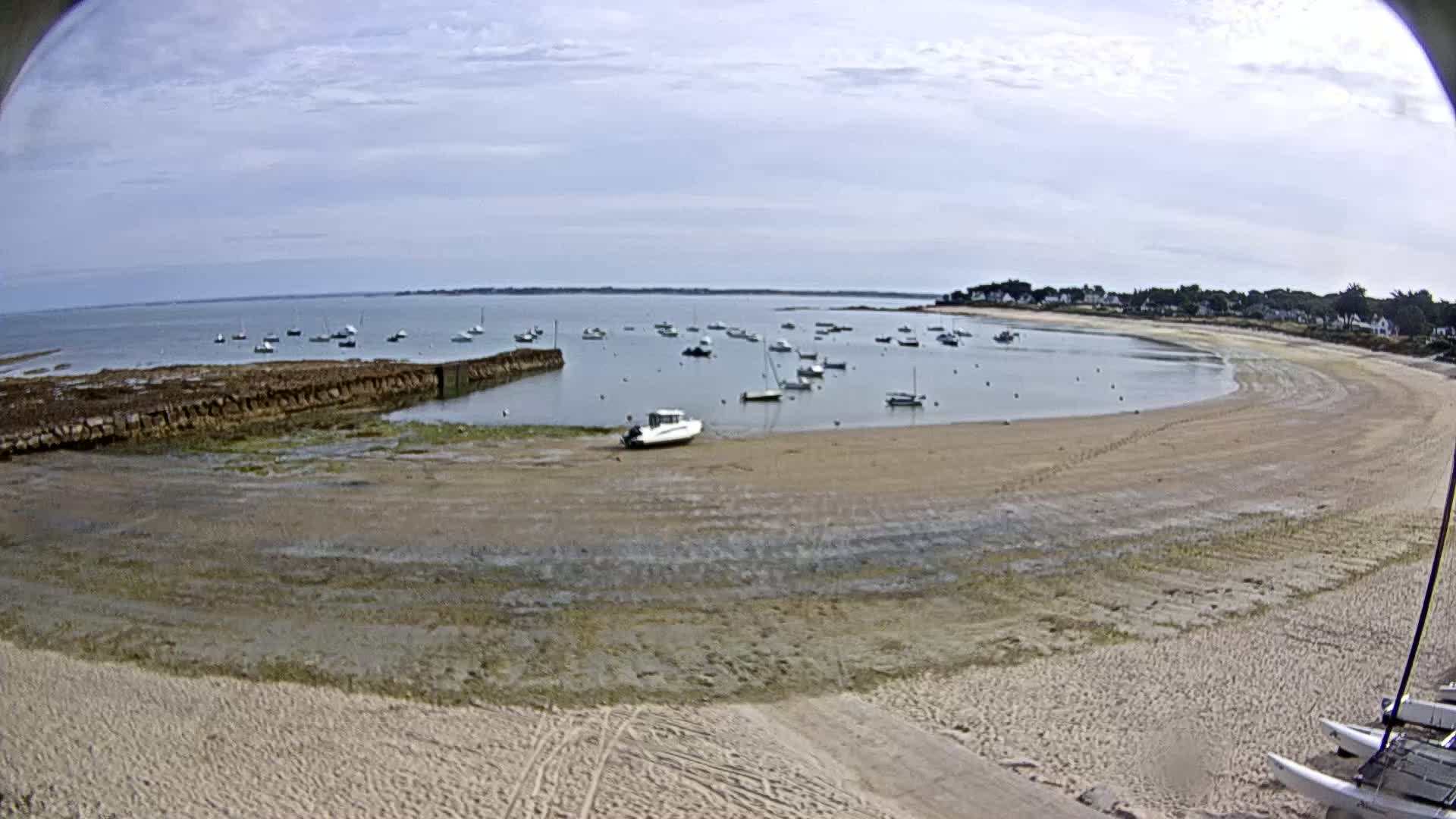 A sandy beach curves around a calm bay containing numerous small boats under a partly cloudy sky.
