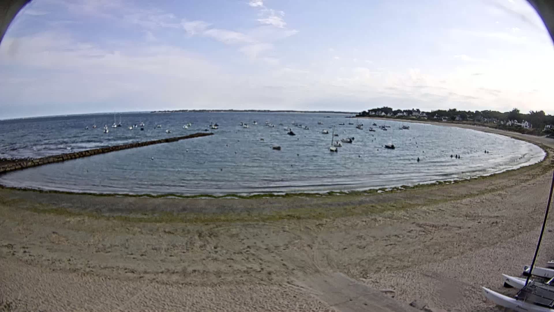 A sandy beach curves around a calm bay filled with numerous small boats under a partly cloudy sky.