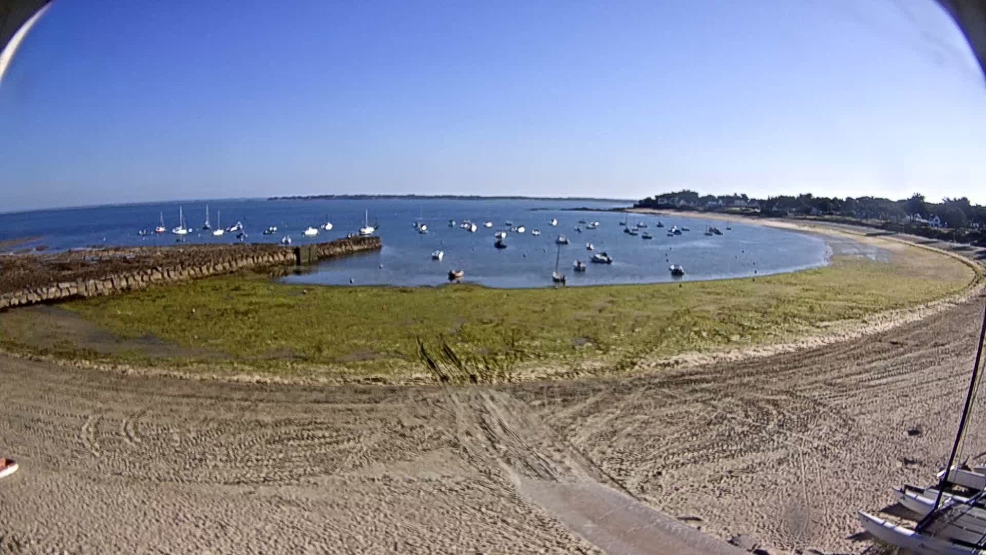 A sandy beach curves around a calm bay filled with sailboats under a clear blue sky.