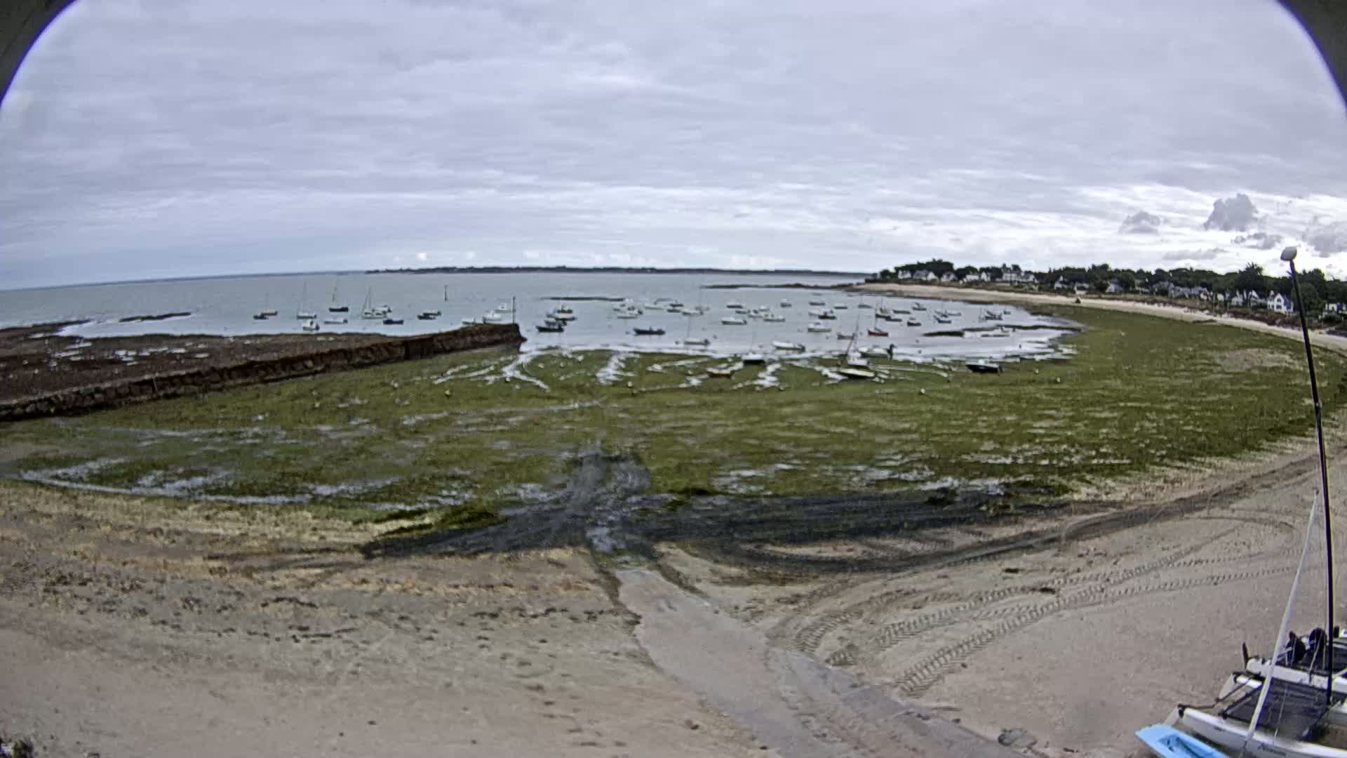 A wide, sandy beach with seaweed-covered mudflats stretches out to a calm bay where numerous small sailboats are anchored under a cloudy sky.