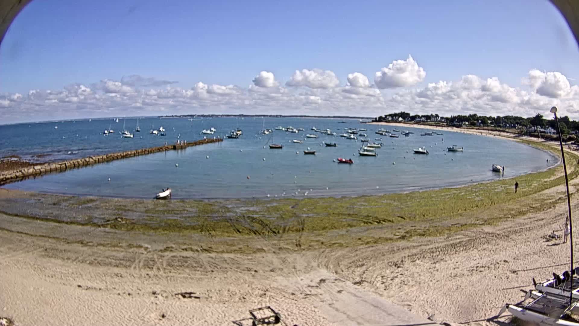 A sandy beach curves around a calm bay filled with numerous small boats under a partly cloudy blue sky.