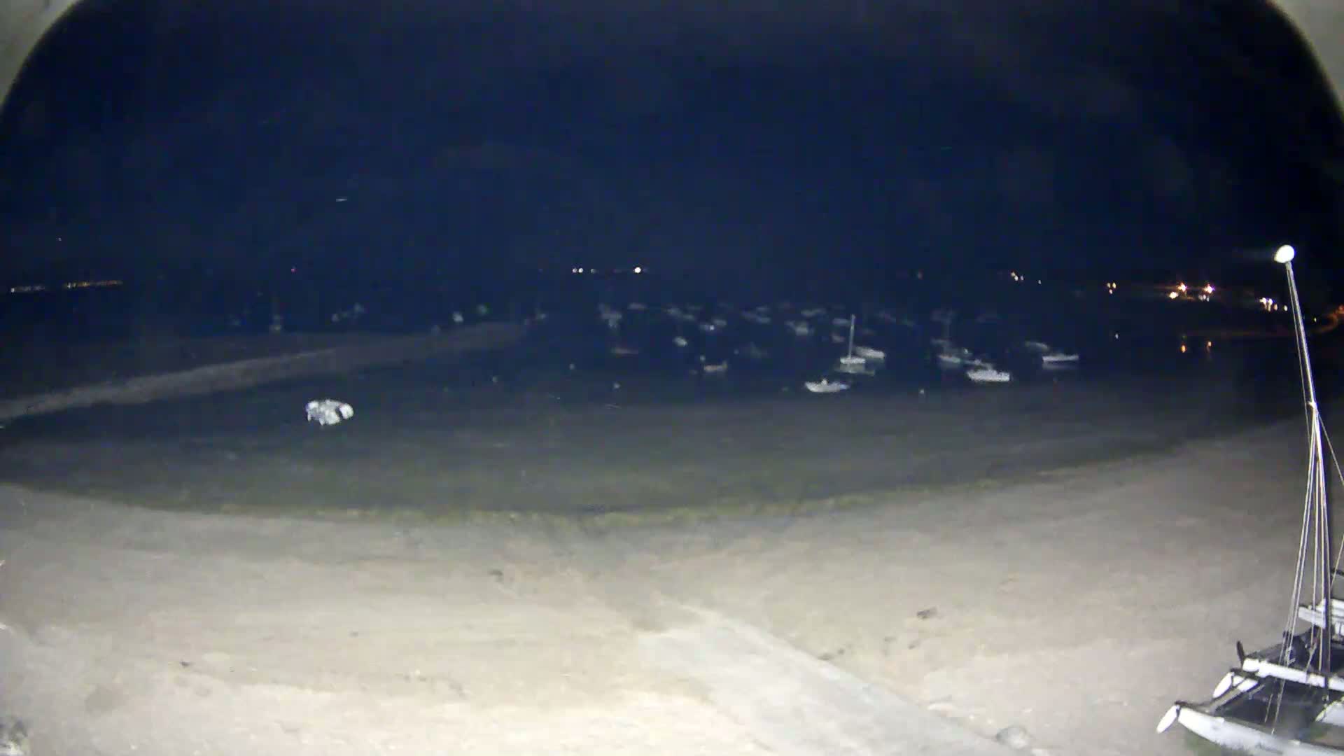 A nighttime view from a slightly elevated position shows a sandy beach, a calm body of water with numerous boats moored, and a distant shoreline with faint lights.