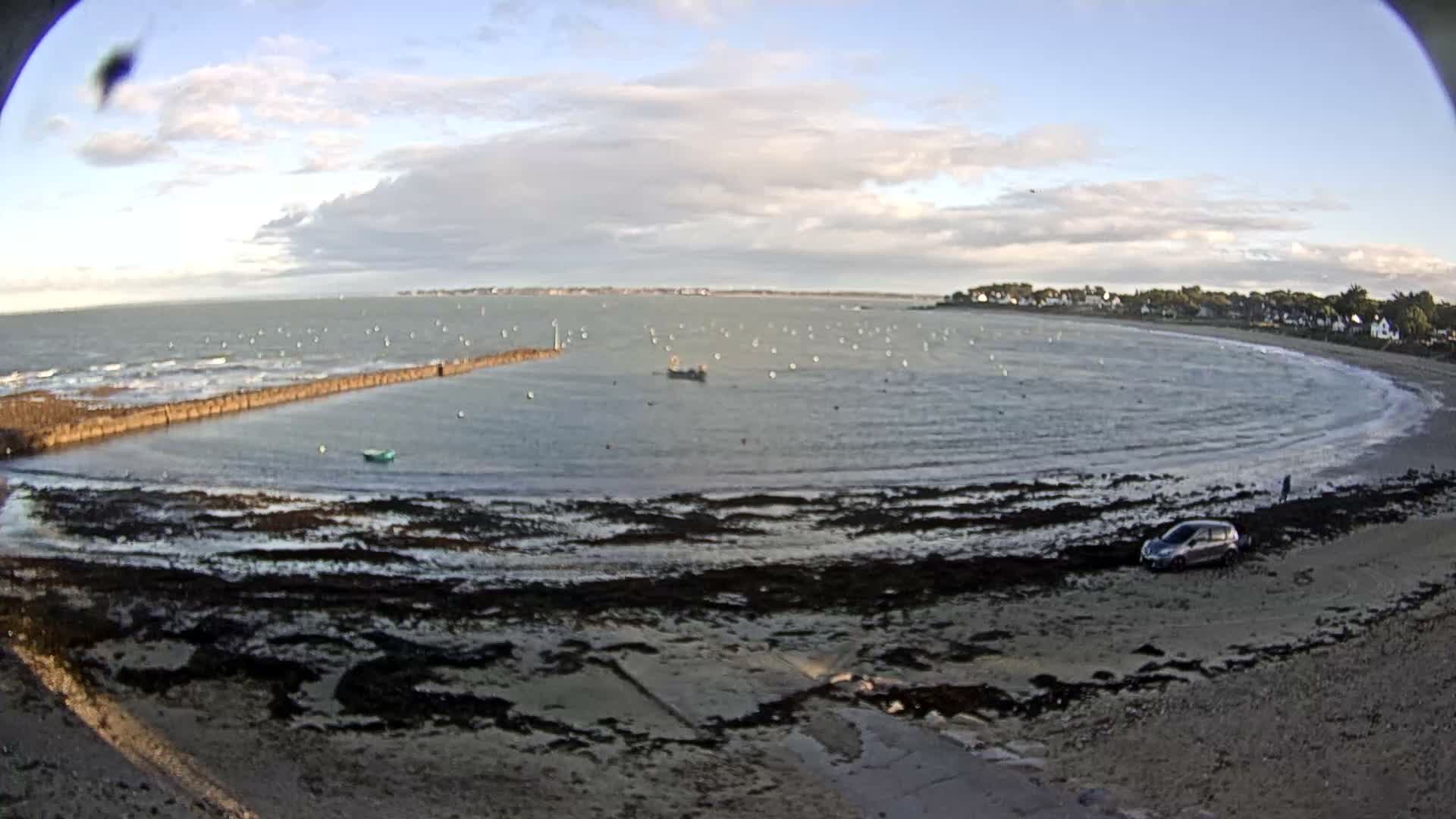 An aerial view captures a wide bay at low tide on a partly cloudy day, featuring a long stone jetty, numerous buoys, a car parked on a seaweed-covered beach, and a distant coastal town lining the shore.