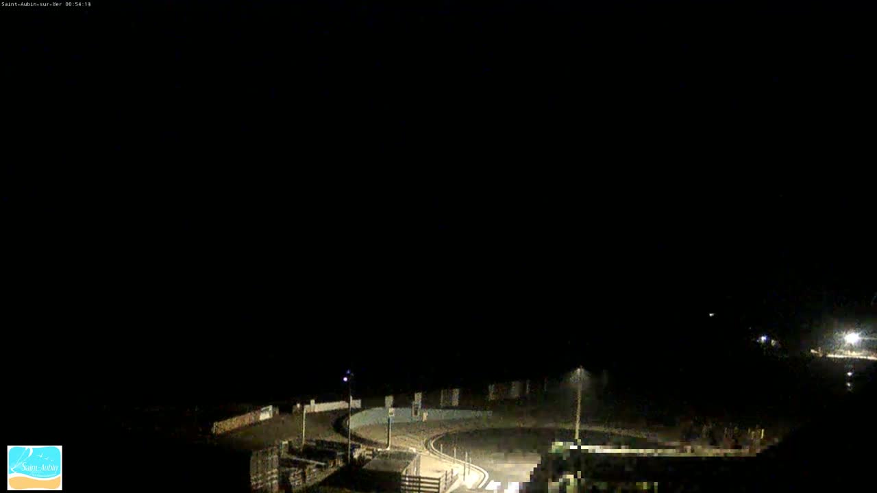 A nighttime, low-light view of a coastal road curving around a dark area, possibly a parking lot or beach, with a few streetlights illuminating the scene.