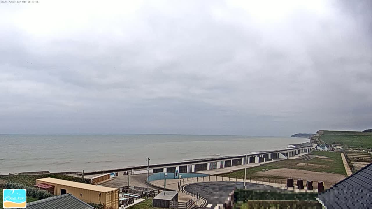 A calm, gray sea stretches to the horizon under an overcast sky, with a long row of beach structures and a grassy area leading up to a cliff in the background.