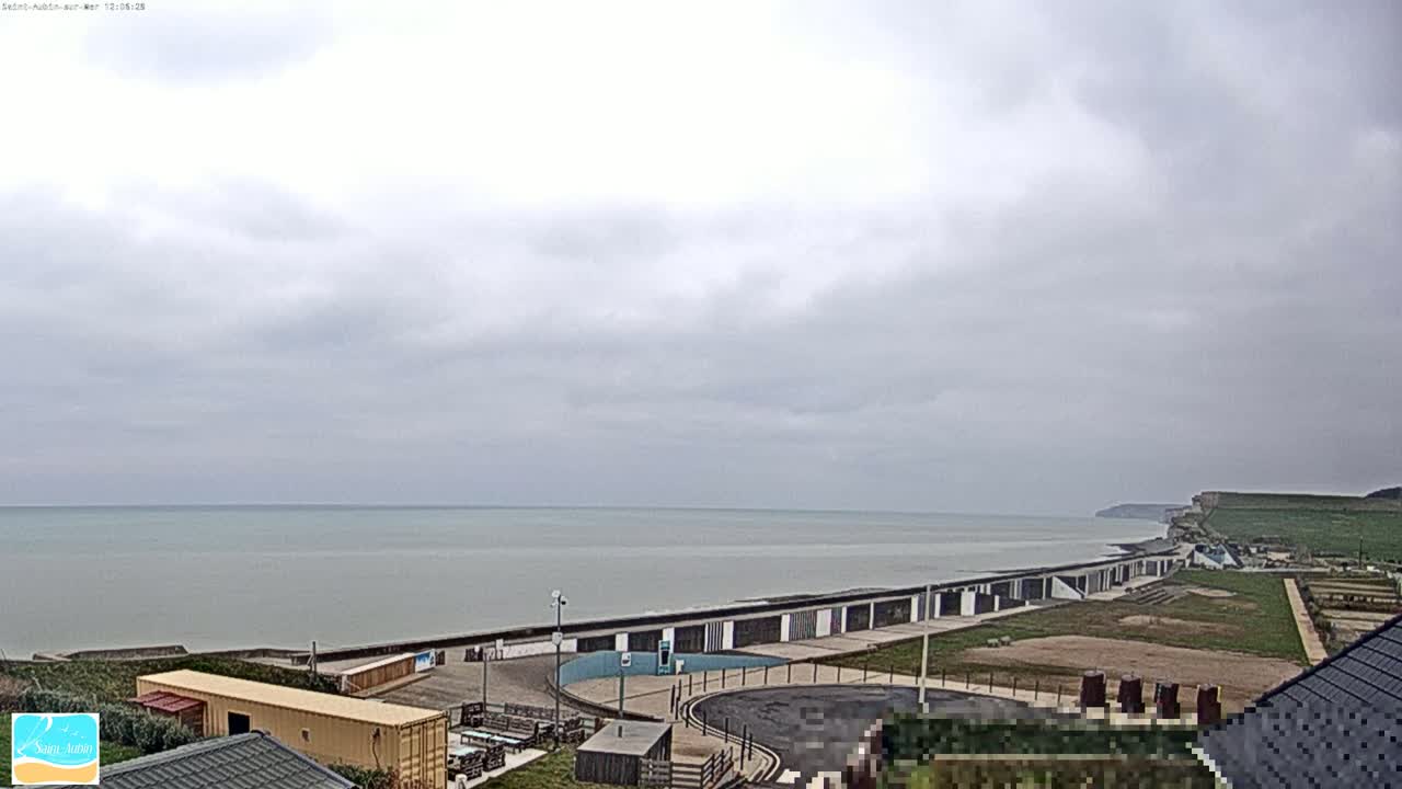 A calm, gray sea stretches to the horizon under an overcast sky, overlooking a coastal area with buildings and a paved walkway.