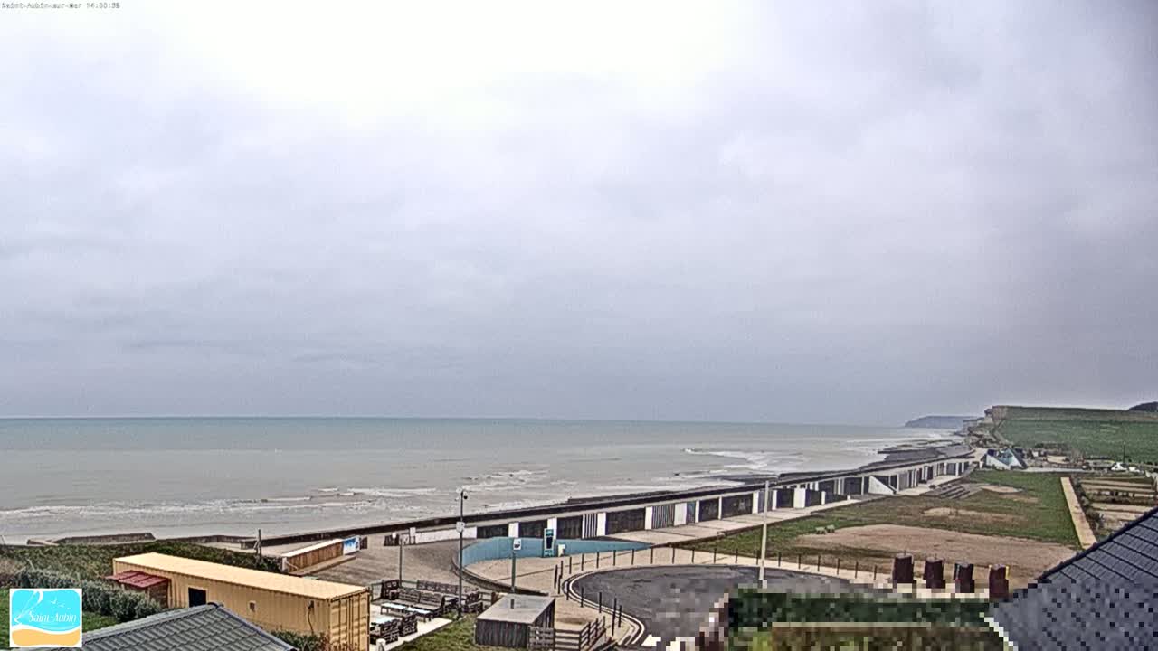 A calm, gray-sky day overlooks a beach with a row of buildings along the shore and grassy areas leading to cliffs in the distance.