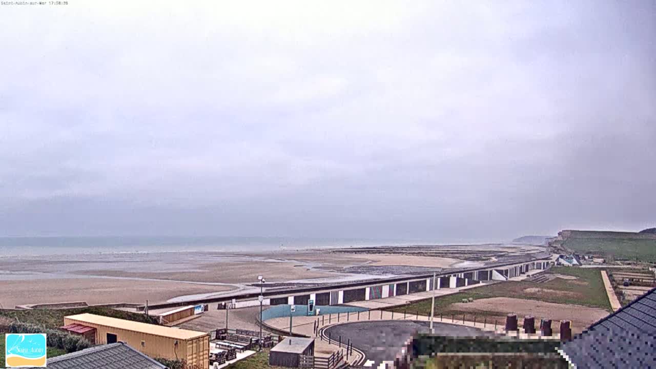 A long, low building complex sits beside a wide, sandy beach under an overcast sky, with a cliff visible in the distance.