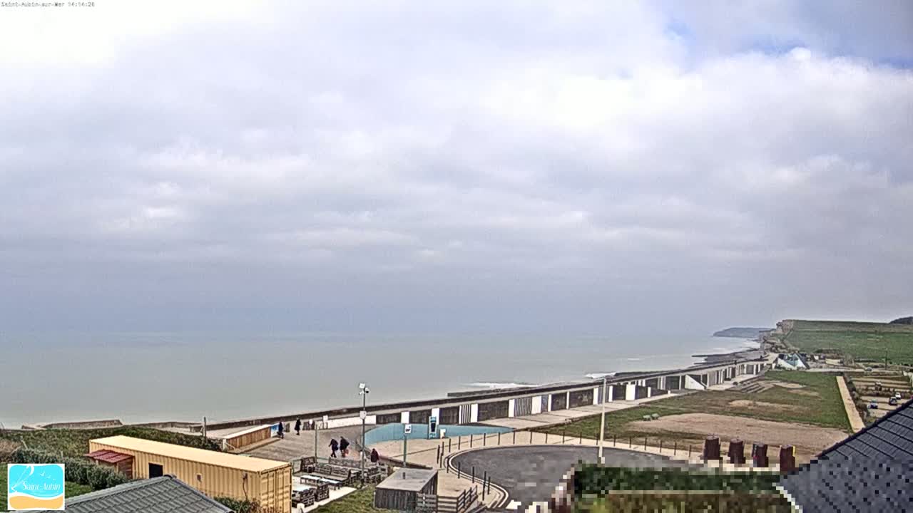 A coastal scene under a mostly cloudy sky shows a long row of beach structures, a paved area, and a grassy hillside leading to a cliff overlooking the calm sea.