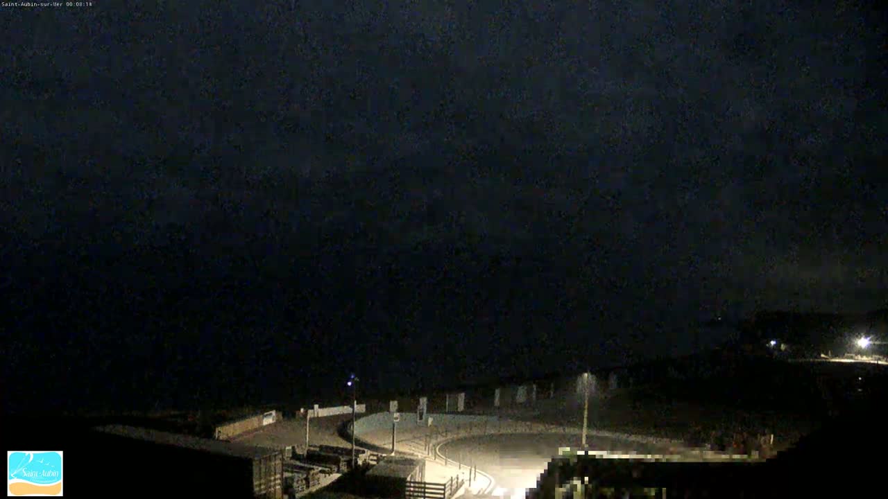 A nighttime, dark overhead view shows a paved, curved roadway overlooking a dark body of water under a cloudy sky.