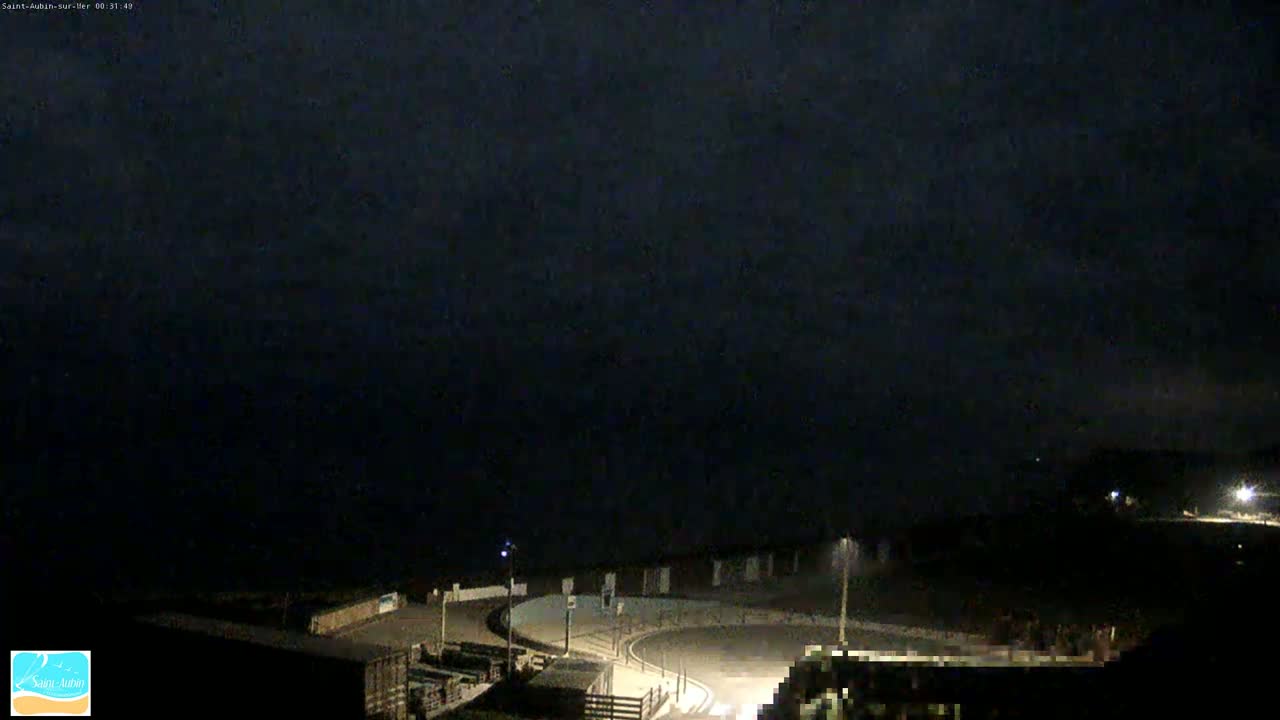 A dark, nighttime scene shows a coastal road curving around a small, dimly lit area near the ocean under a mostly cloudy sky.