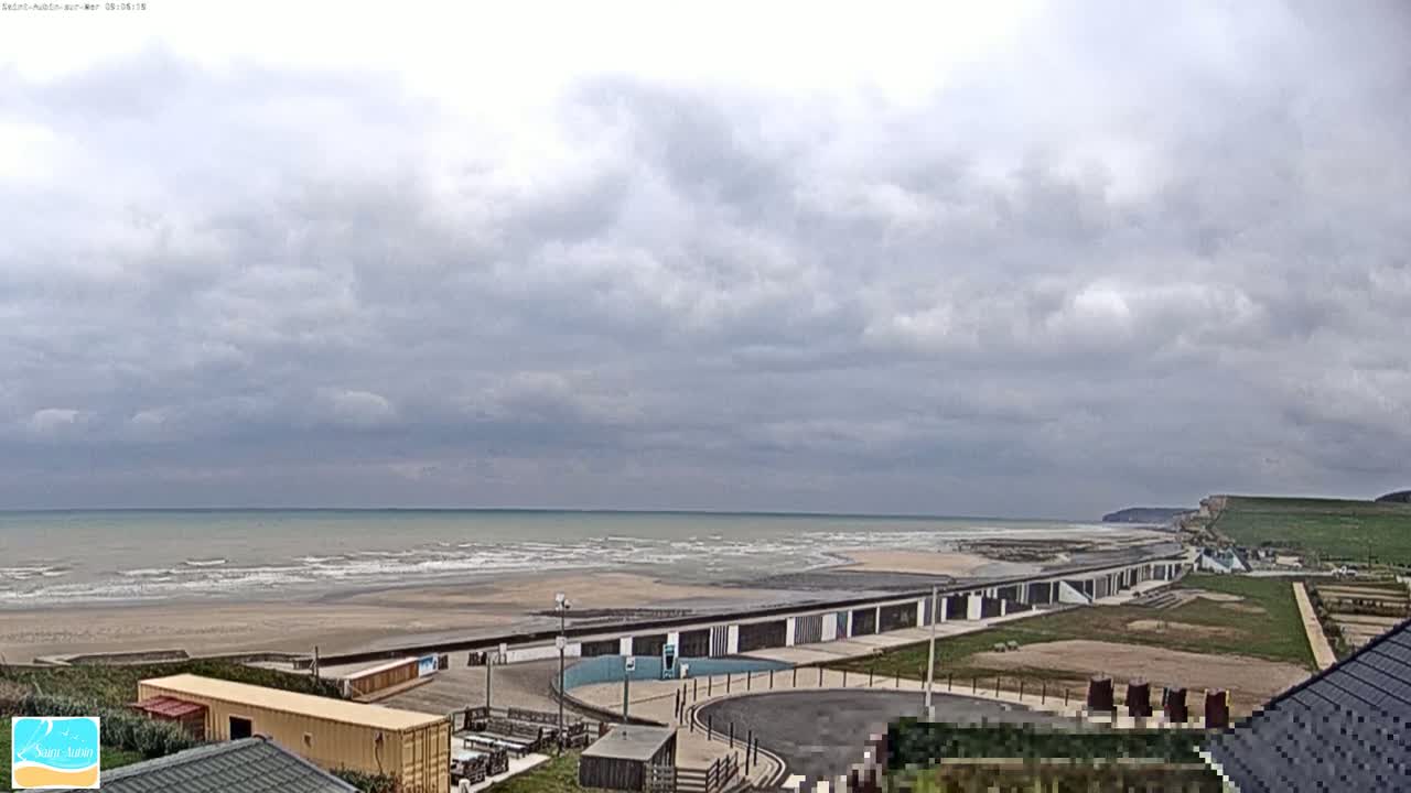 A coastal scene under a cloudy sky shows a wide sandy beach with gentle waves, beachfront buildings, and a grassy area leading up to a cliff in the distance.