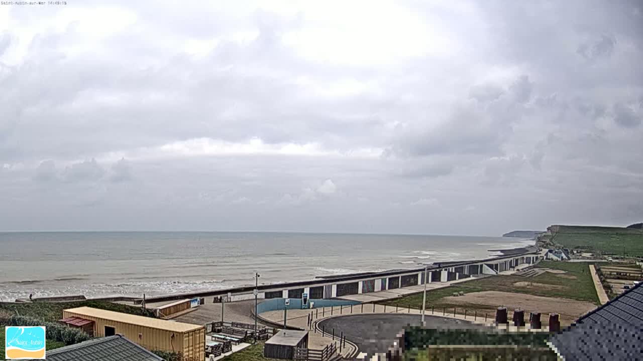 A mostly cloudy day overlooks a calm ocean meeting a shoreline with buildings and grassy areas.