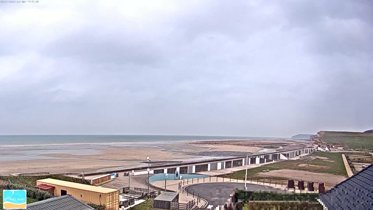 A wide shot shows a beach with a long, low building along its edge, under an overcast sky.