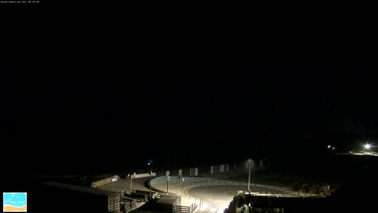 A nighttime, overhead view of a dark, empty roadway curving around a structure, with only a few lights visible.