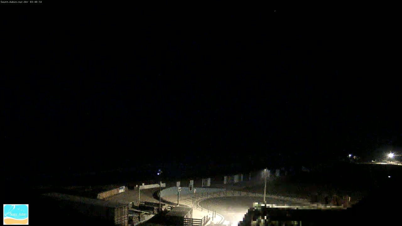 A nighttime, dark view of a paved roadway curving around a small, dimly lit area near the ocean under a clear sky.