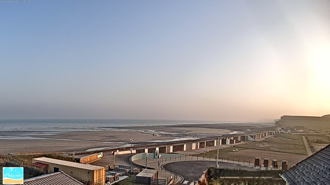 A long sandy beach stretches to a calm ocean under a clear, pale-blue sky at sunset, with beach huts and buildings visible in the foreground.